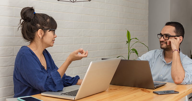 cheerful-loving-young-latin-couple-using-laptops-talking-working-home-sitting-long-table-using-laptops-home-office-concept